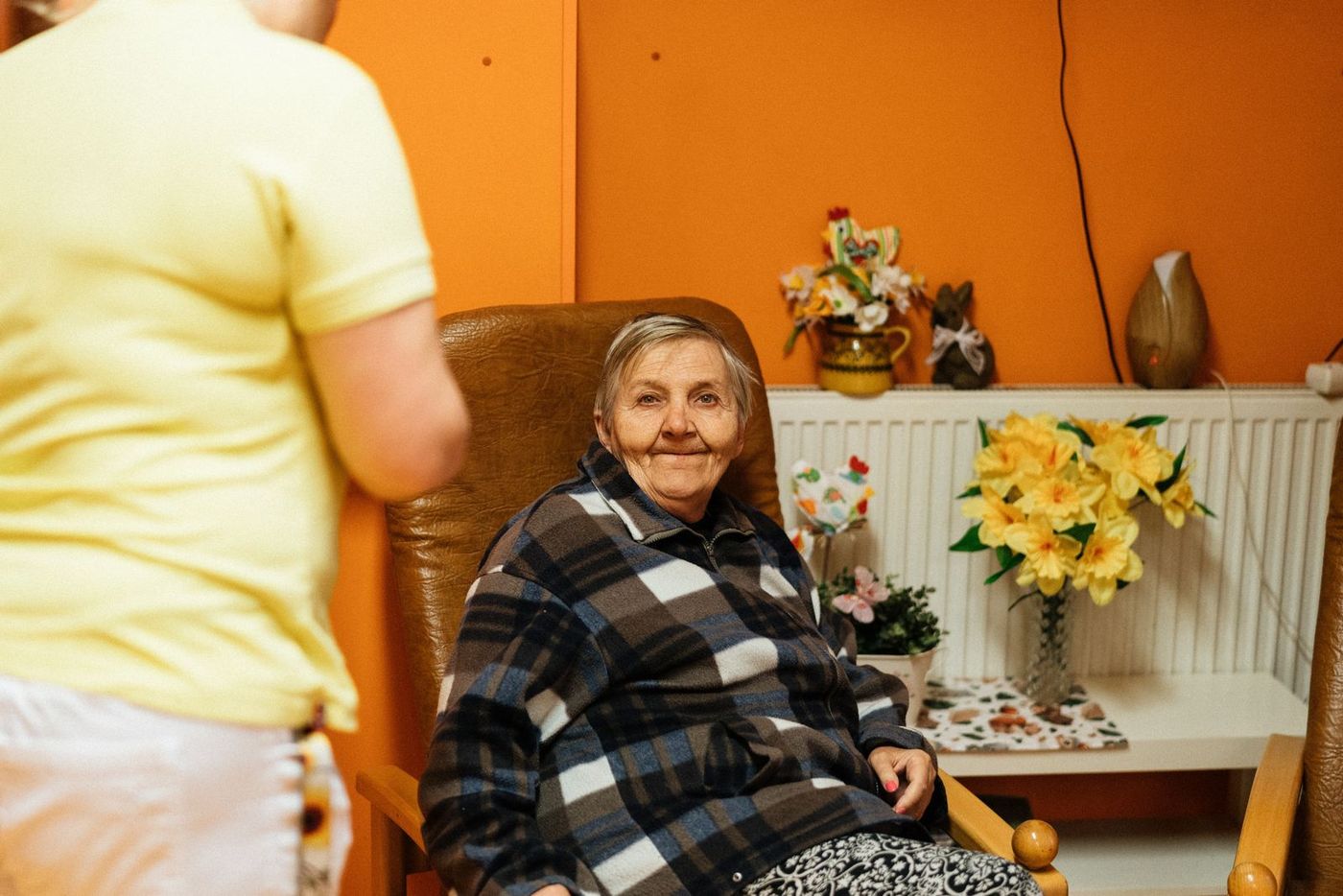 An older woman sitting comfortably in an armchair at home with a carer nearby